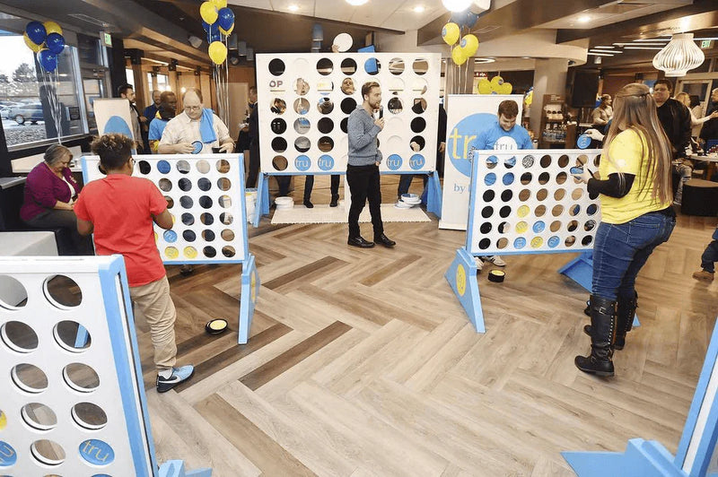 A diverse group of people playing a giant wooden Connect Four game in a lush green park, surrounded by other oversized games like a giant chess set and a tumble tower.