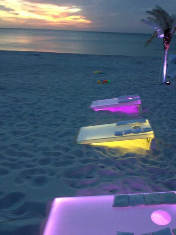 An outdoor nighttime event featuring a well-lit cornhole game with LED lights, surrounded by oversized chess pieces and other giant outdoor games in a picturesque garden setting with event organizers engaging in the activities.